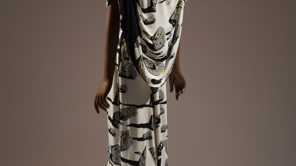 Long evening dress in ivory rayon crinkle crepe printed with surrealist design of gray shattered monument stones with black shadows and yellow ancient inscriptions of keys, insects, eyes, timepieces, words, arrows, birds; deep-V suplice bodice with cap sleeves, asymmetrical black draped capelet, flared train