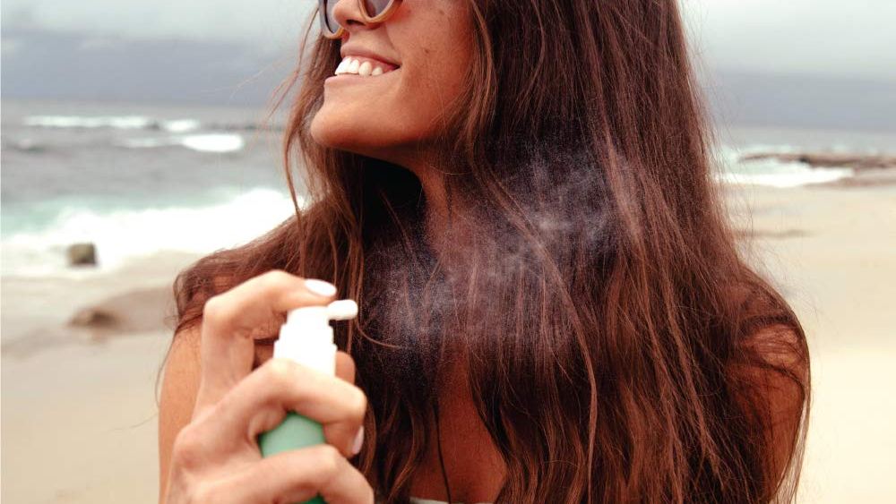 A woman applying the COOLA Organic Scalp & Hair Sunscreen Mist at the beach