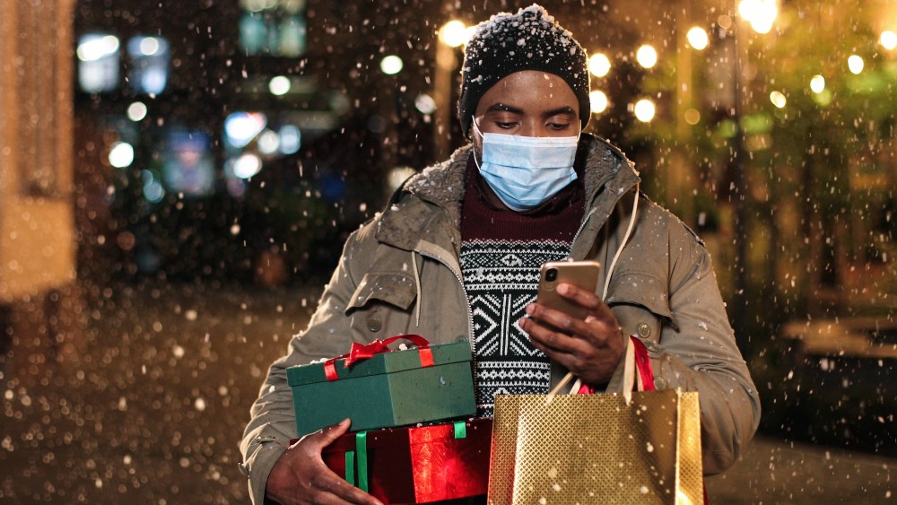 Portrait of man standing and texting on mobile phone. Young African American happy male with little Christmas tree and shopping bags typing on smartphone while snowing. Winter holidays concept