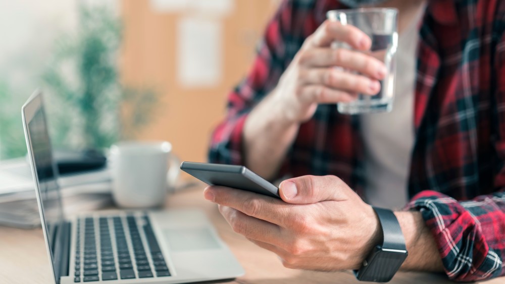 Gig worker drinking water and using mobile phone in home office during freelance project work. (Photo by: SCIENCE PHOTO LIBRARY via AP Images)