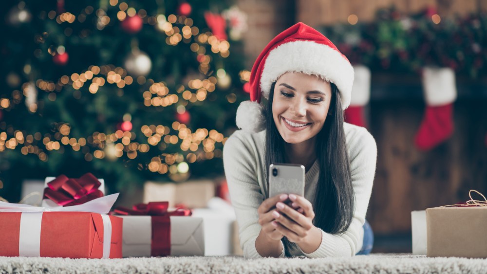 Close up photo of positive girl in santa claus hat lie on carpet use smartphone send christmas, congratulation search discounts in house with x-mas comfort atmosphere indoors