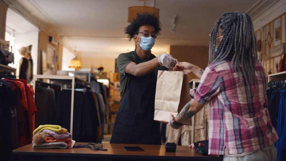 Young woman in safety mask purchasing in clothes store. African american shopkeeper man wearing medical mask and gloves working at clothing store