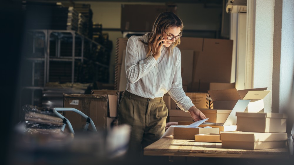 Woman online seller confirming orders from customer on the phone. E-commerce business owner looking at the papers and talking on phone in store warehouse.