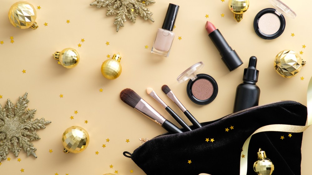 Makeup bag with cosmetic products and golden Christmas decorations spilling out on to pastel beige background. Glamour make up artist pouch with professional beauty products