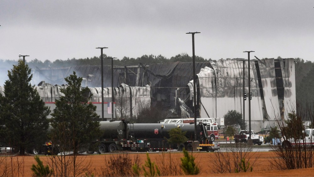 Smoke continues to rise from gutted areas of the QVC shipping facility in Rocky Mount, N.C. Sunday morning, Dec. 19, 2021, as firefighters continue to spray water into hot spots. Much of the building was severely damaged by a fire that started early Saturday morning. Water tankers are visible in the foreground.  (Scott Sharpe/The News & Observer via AP)