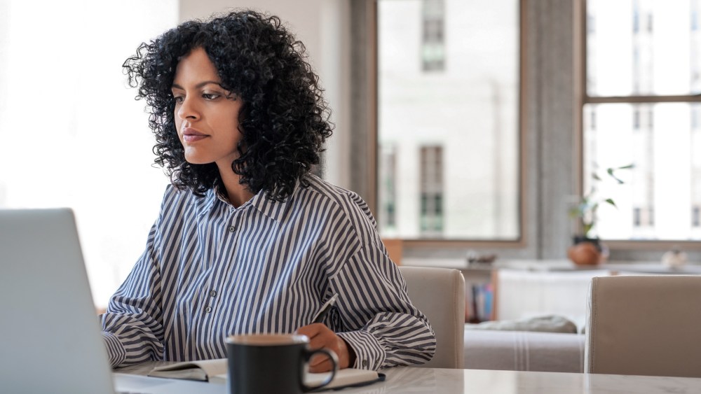 A women with curly dark hair in a striped button-up shirt working from her laptop in her sunny apartment with coffee on the table.