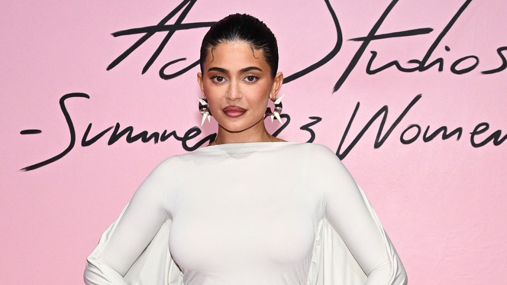 Kylie Jenner attends the Acne Studios Womenswear spring 2023 show at Paris Fashion Week on Sept. 28, 2022 wearing white cape dress and standing in front of pink background.