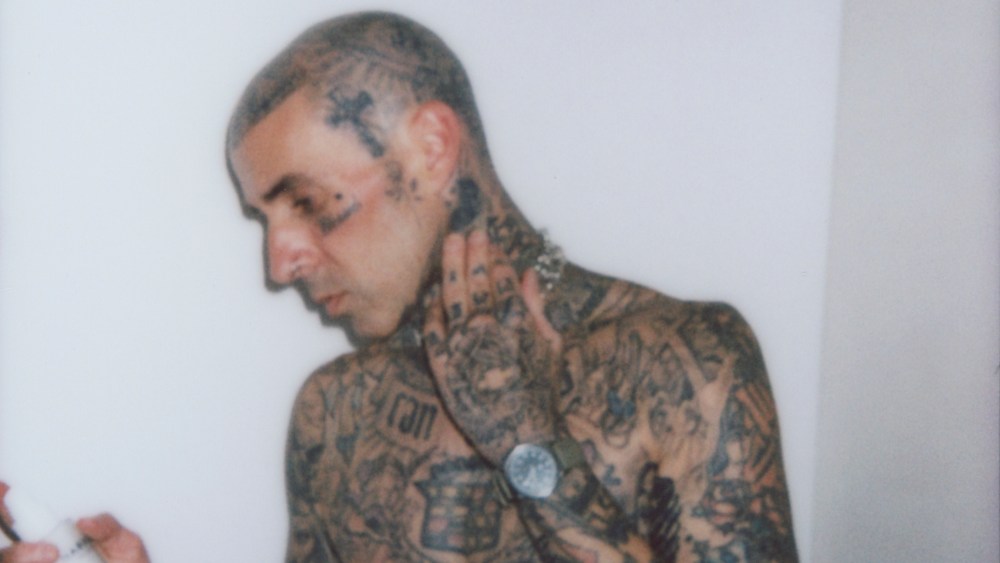 Travis Barker shirtless displaying his full body tattoos and holding a bottle of from his skin care line.