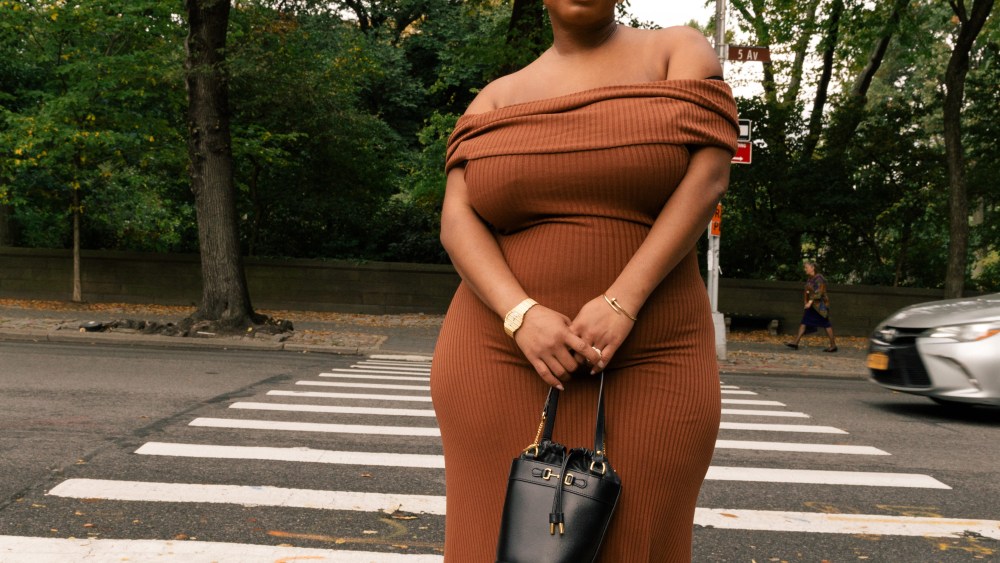 A model with sunglasses and shaved head for The RealReal wearing a plus-size auburn bodycon dress and posing uptown.