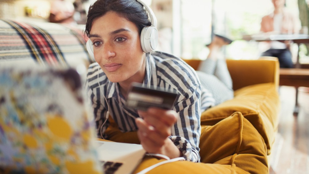A young woman with headphones on peruses the web with her credit card in hand, reclining on a mustard yellow sofa.