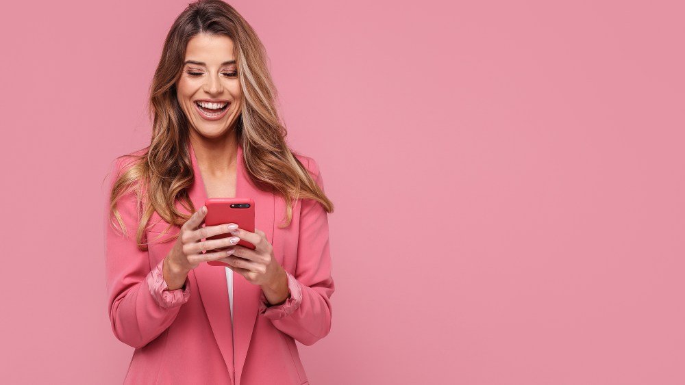 Photo of a happy young beautiful woman using mobile phone, smiling, posing isolated over pink pastel studio background.
