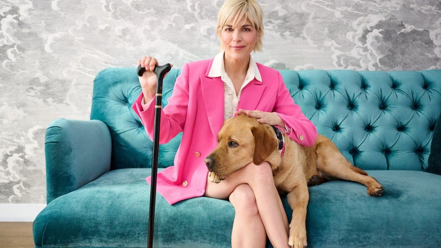 Selma Blair, QVC Brand Ambassador for Accessibility, sits on a plush teal sofa with her dog, Scout. She wears a hot pink suit and uses a black and gold cane.