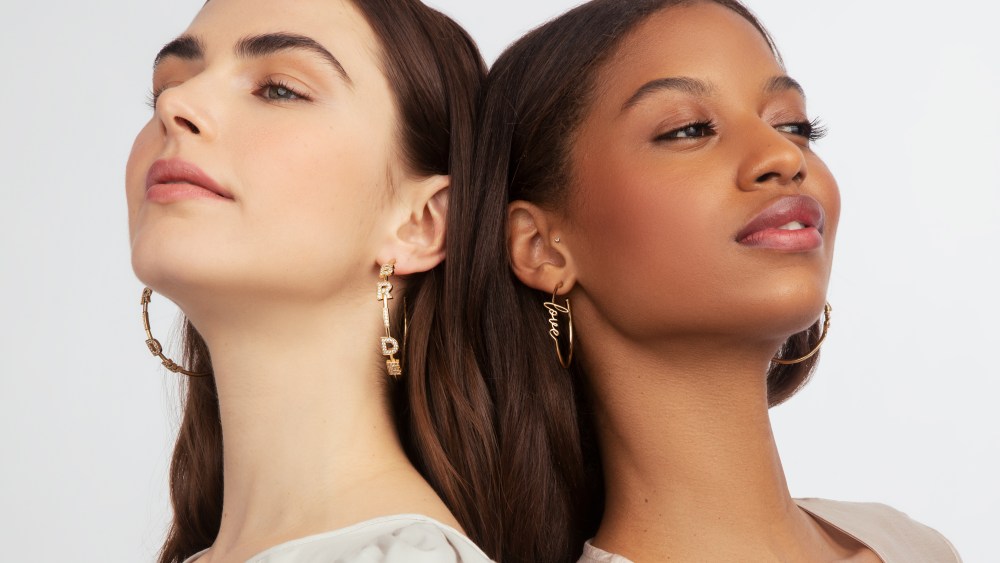 Two women stand back to back with their heads resting on each other, each wearing earrings from the Kleinfeld jewelry collection. One hoop earring says "Bride" and another hoop earring says "love."