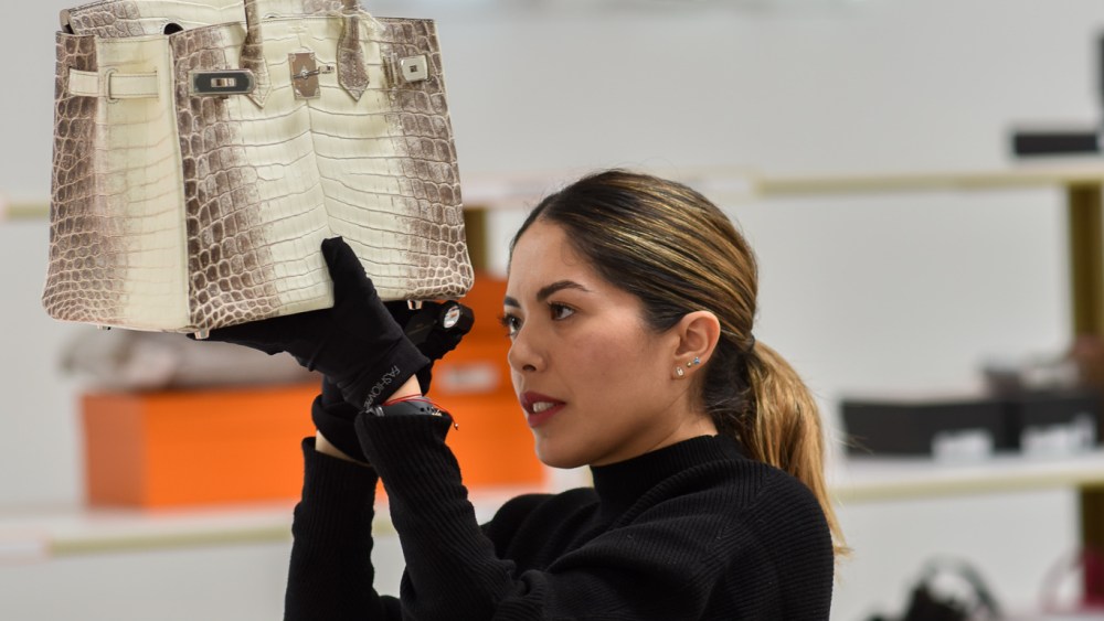 Senior authentication manager at Fashionphile Laura Chavez inspects a cream crocodile Hermes bag in front of a neon sign that reads "Authentication Station."