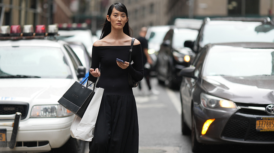 A model wears a black off-shoulder top with long sleeves, a black long skirt, black leather shoes, a shopping bag, outside Jason Wu, during New York Fashion Week, on September 10, 2023 in New York City.