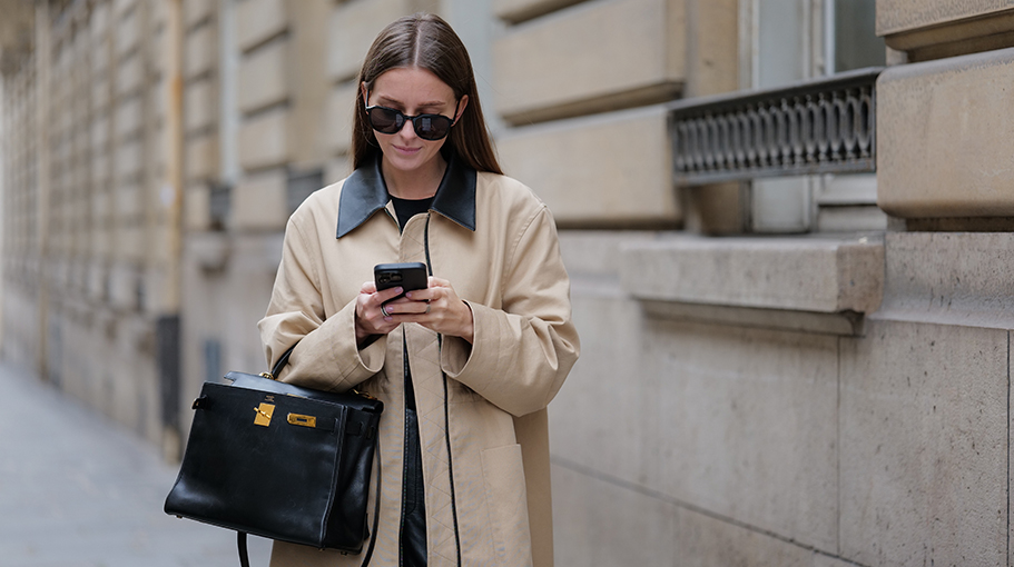 Diane Batoukina wears sunglasses, a beige trench coat with black collar, a black t-shirt, a black leather Hermes bag, black leather shiny pants, during a street style fashion photo session, on March 17, 2023 in Paris, France.