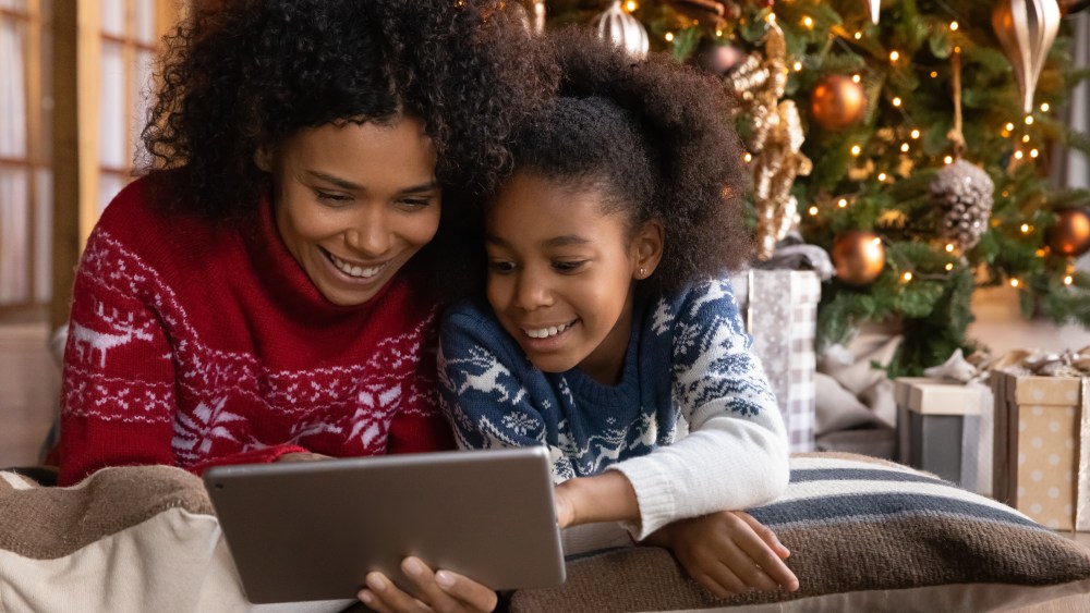Close up happy African American woman with daughter using tablet together, lying on soft pillows near Christmas tree at home, happy family shopping online, choosing gifts, enjoying winter holiday
