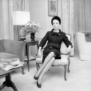 Outtake; Gloria Guinness seated in her suite in the Waldorf Towers wearing a black wool Givenchy jacket bordered in black faille over a sleeveless dress with a black Balenciaga hat and  diamond ring and lapel pin as well as pearls and diamond and pearl earrings; black round-toe pumps  completed the dinner outfit (Guinness owned the exact same outifts in mauve and blue linen with a beige hat)..Article title: 'Mrs. Loel Guinness'