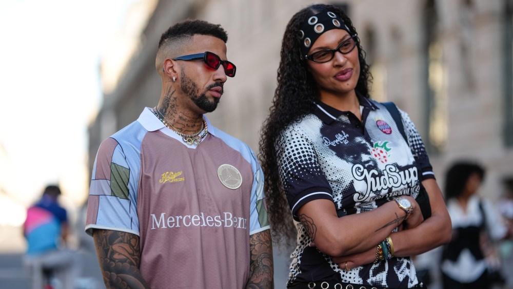 Elizabeth Cambage wears black silver circle headband, black sunglasses, silver necklace, black white graphic pattern/print KidSuper x Ronaldinho sports jersey shirt, black silver circle mini shorts, shiny black leather bag. Miles Chamley-Watson wears a Kid Super x Mercedes-Benz short-sleeved shirt featuring a geometric Mercedes-Benz design in shades of pink, white, and light blue, outside KidSuper, during Paris Fashion Week - Menswear Spring/Summer 2026