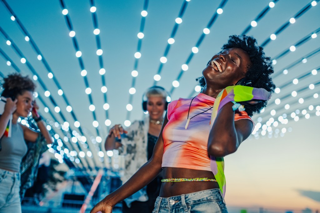 Happy friends wearing headphones dance joyfully together at a vibrant silent disco, surrounded by colorful decorative lights, celebrating friendship and carefree moments under the stars