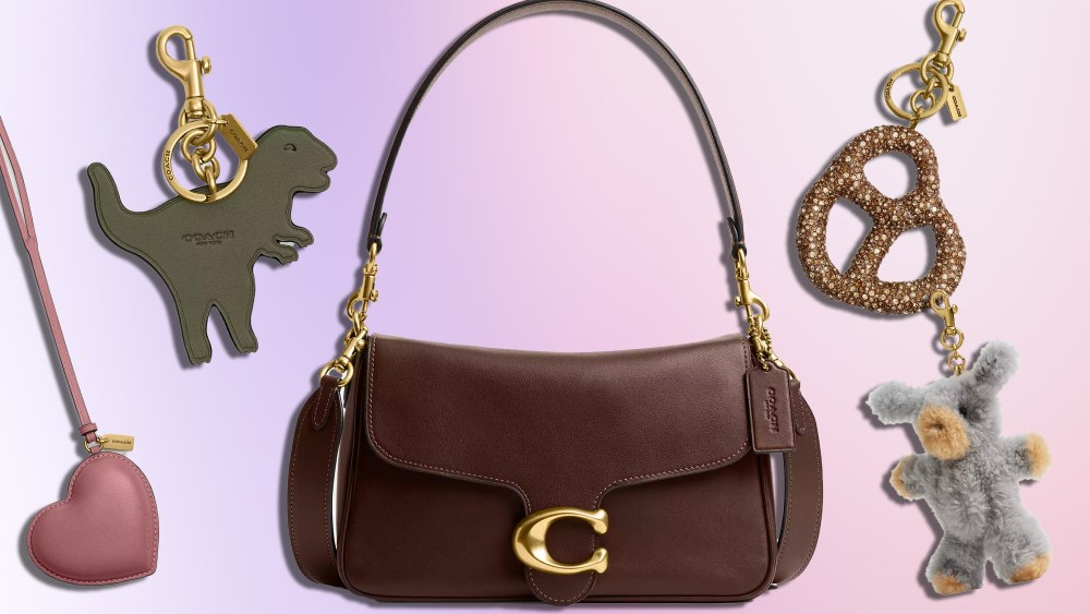 brown soft tabby bag from coach positioned next to an assortment of bag charms, including a pretzel, teddy bear, heart, and dino over pink gradient background