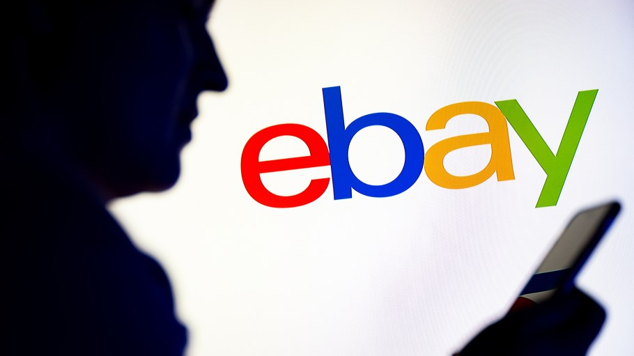 In this photo illustration, a silhouetted individual is seen holding a mobile phone with the eBay logo displayed in the background.