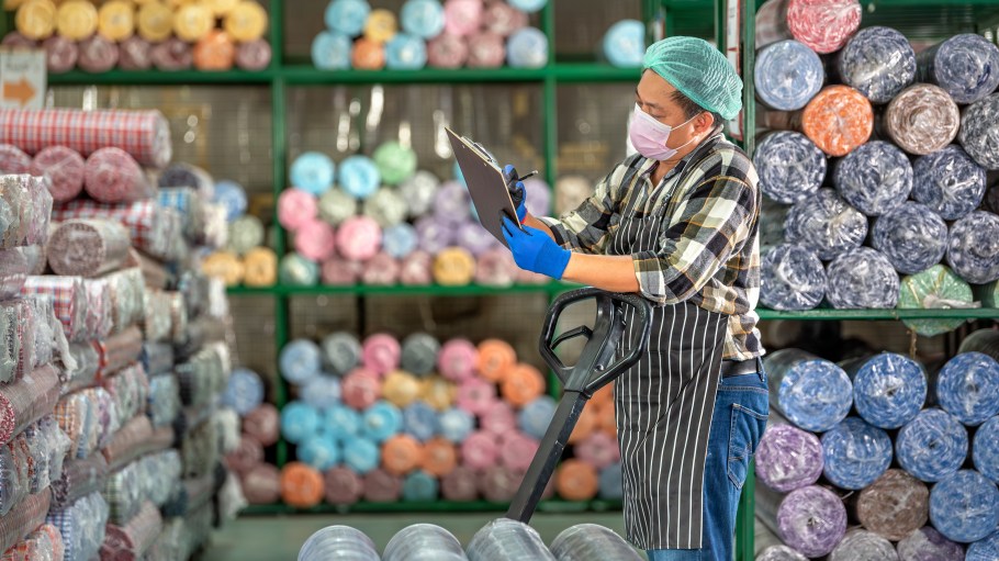 Warehouse worker in mask and gloves standing with pallet Jack holding clipboard and checking inventory of colorful fabric rolls on shelves. Male staff auditing textile stock in storage warehouse.